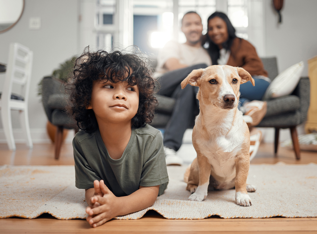child smiling at their dog while laying on a living room floor, with parents in the background