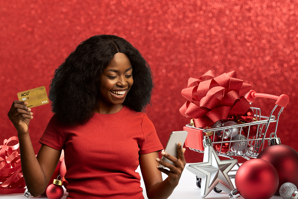Smilling woman looks at her phone while holding a credit card. She's posed next to a box of festive red decorations.