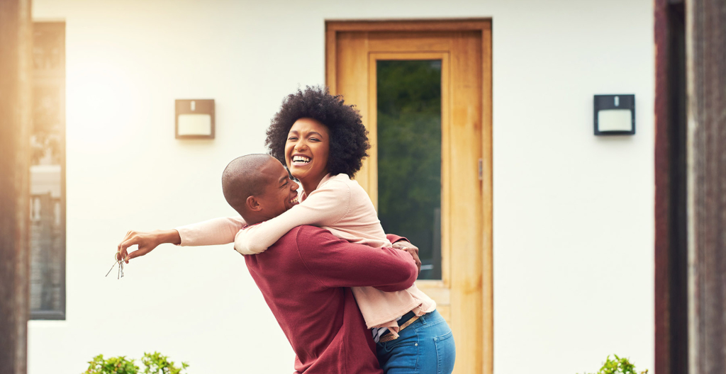 A couple embraces in front of a home, laughing happily and holding keys