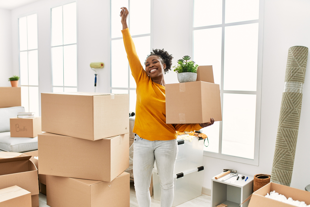 A woman stands in a home or condo holding a moving box, smiling brightly. Other boxes surround her.