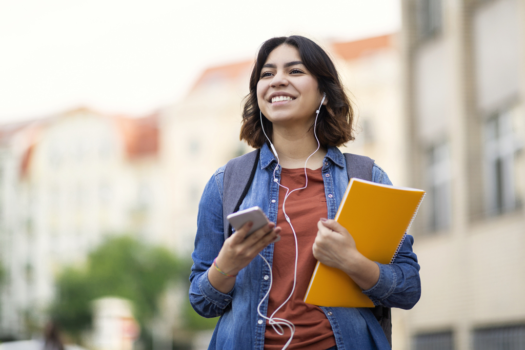 Smiling student on campus,  holding a notebook and phone, wearing wired ear buds