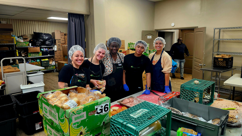 Volunteers smile in a kitchen, surrounded by boxes of food