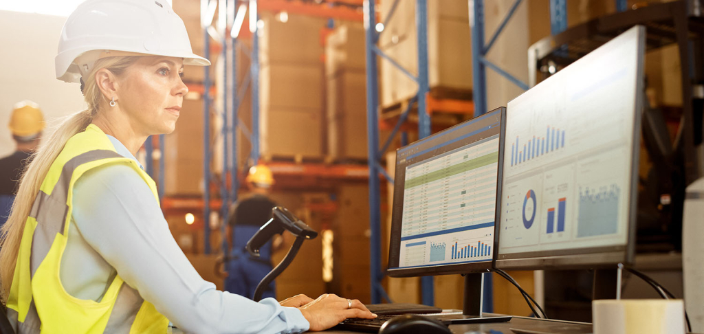 Female business owner wearing hard hat Wworks on Ppersonal computer counting stock in the retail warehouse full of shelves with goods. Commerce, Distribution, Logistics, cash flow