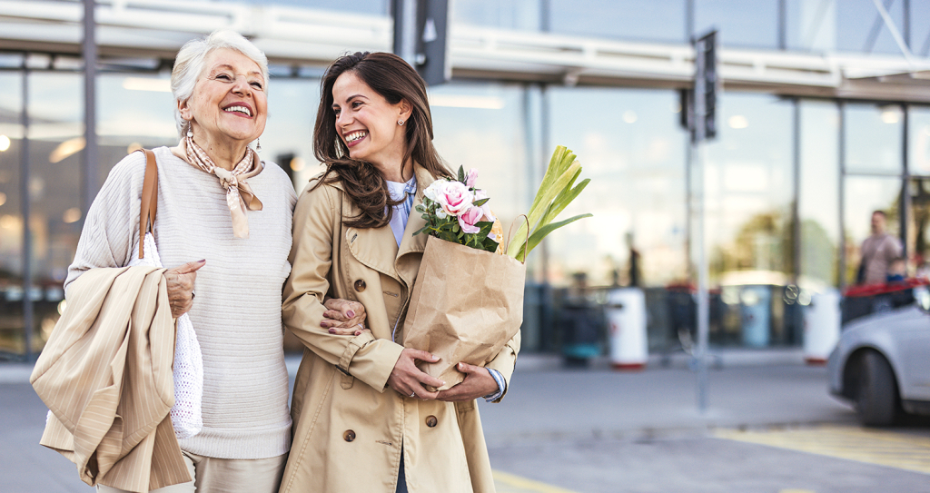 An adult daughter walks with her elderly mother outside a store, carrying grocery bags and sharing a light-hearted moment together.