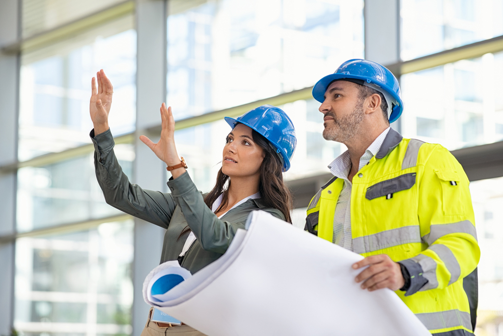 Young woman architect explaining blueprint to supervisor wearing safety vest at construction site. Mid adult contractor holding blueprint and understanding manager vision at construction site. Smiling engineer with hardhat on head talking to contractor while standing in building in construction process.