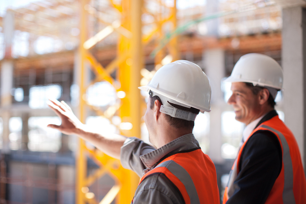 Two men in safety vests and hard hats survey a construction site.